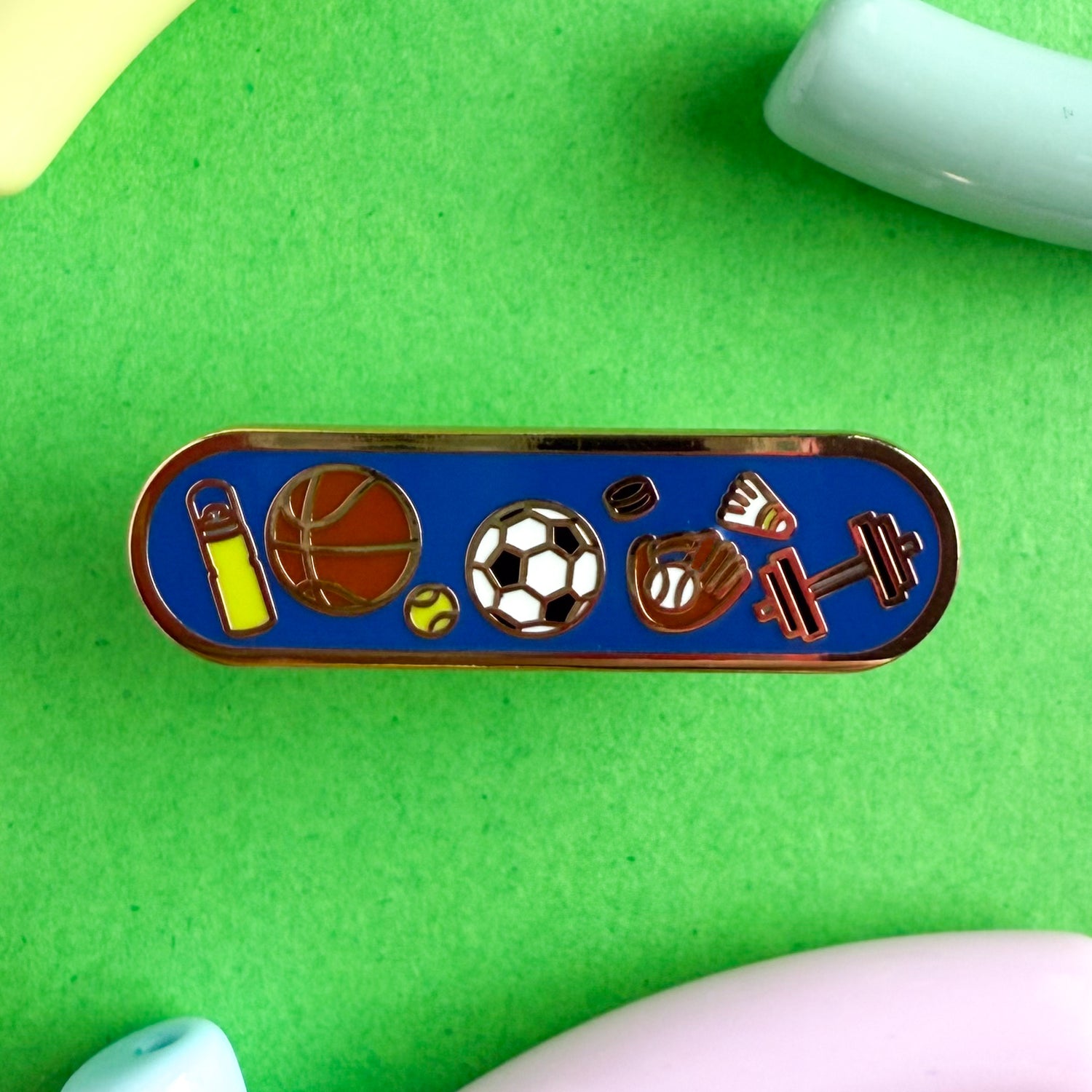 A bandaid shaped enamel pin with sport themed illustration. The pin is on a green background.