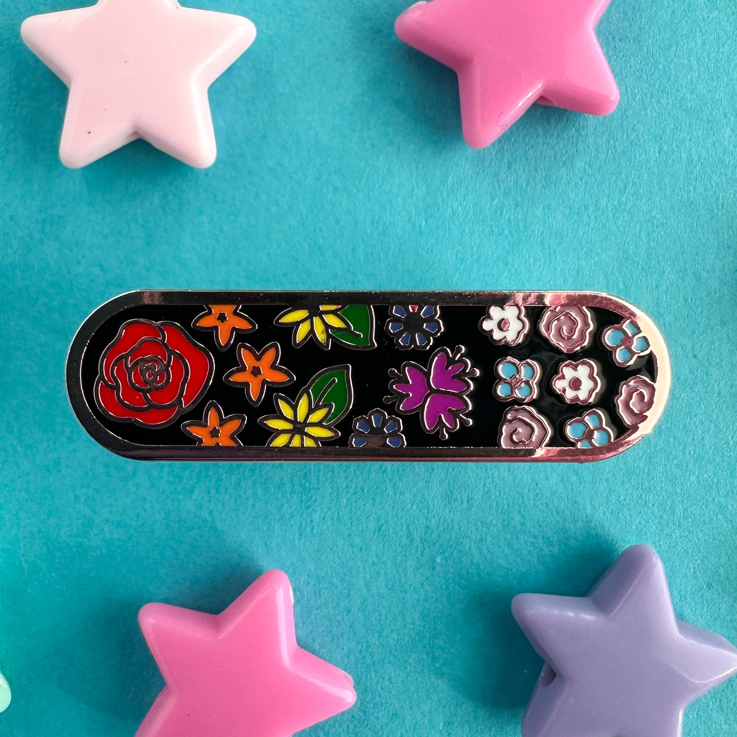 An oval shaped pin with flowers on it in the colors of the rainbow and trans pride flags. The pin is on a blue backdrop with plastic star beads around it. 