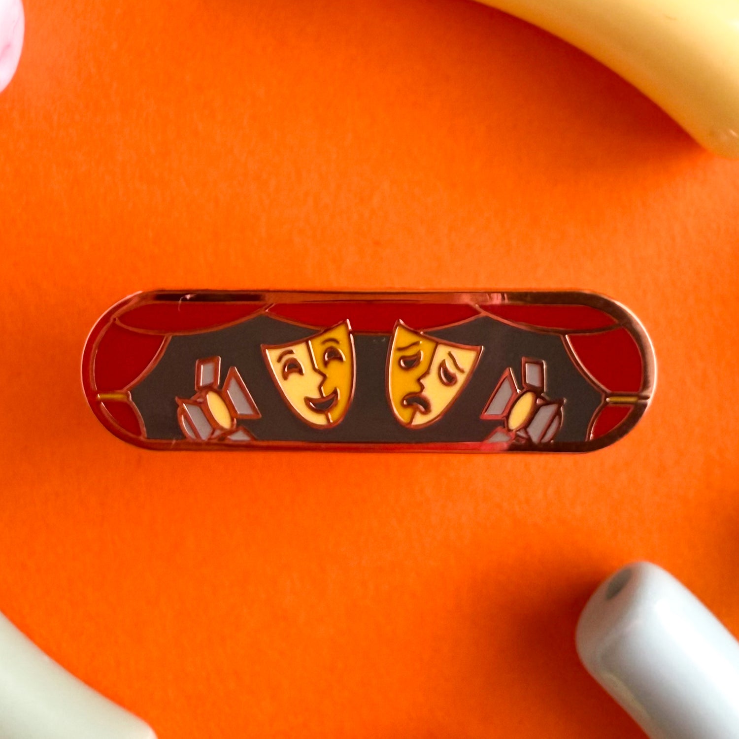 A bandaid shaped enamel pin with theater themed illustrations. The pin is on an orange background.