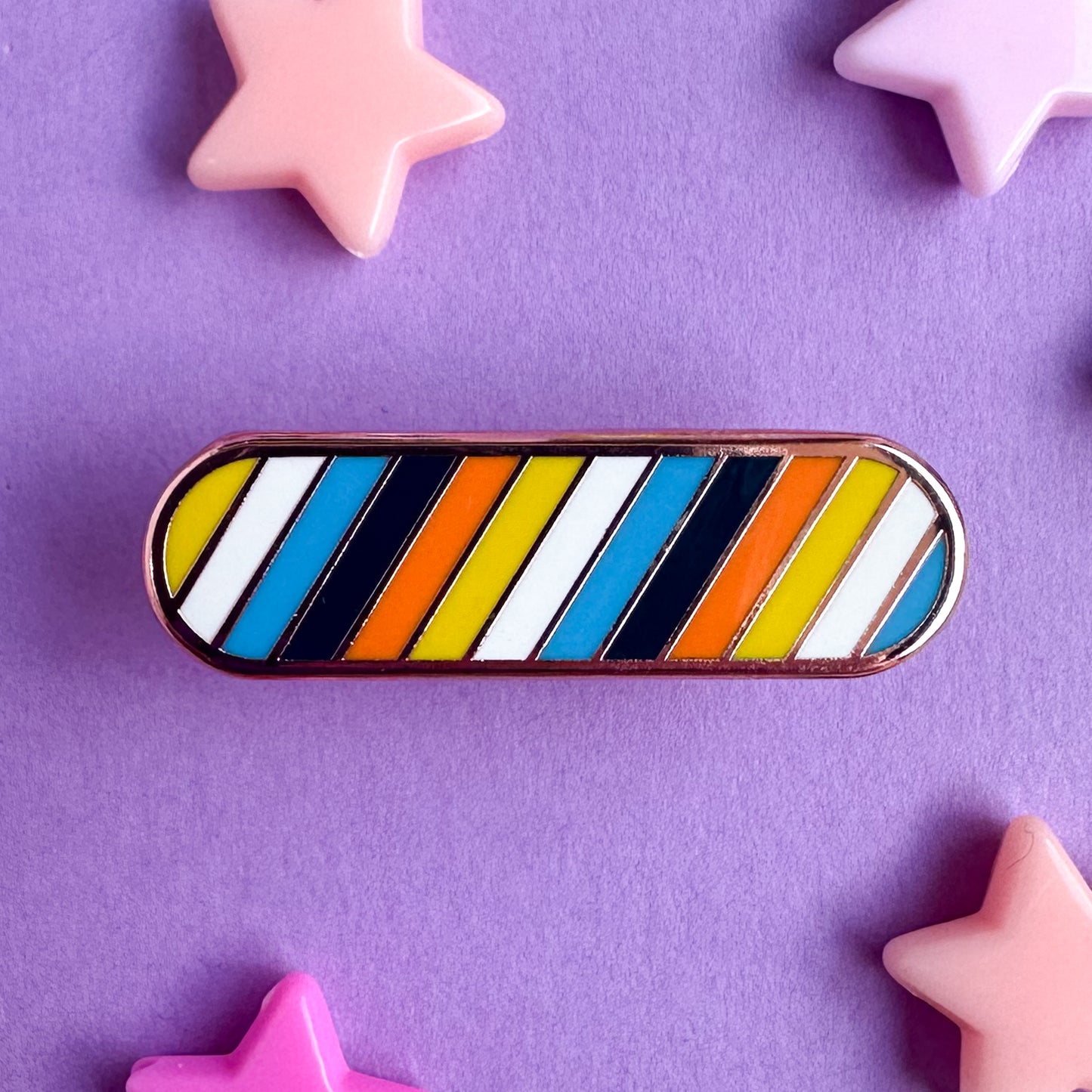 An enamel pin in the shape of an oval with diagonal stripes in the colors of the Aroace Pride flag. The pin is on a purple background with star beads around it. 