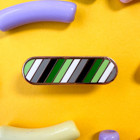 A bandaid shaped pin with diagonal bands of color in the colors of the Aromantic Pride Flag, green, light green, white, grey and black. The pin is on a yellow background with macaroni shaped beads around it.