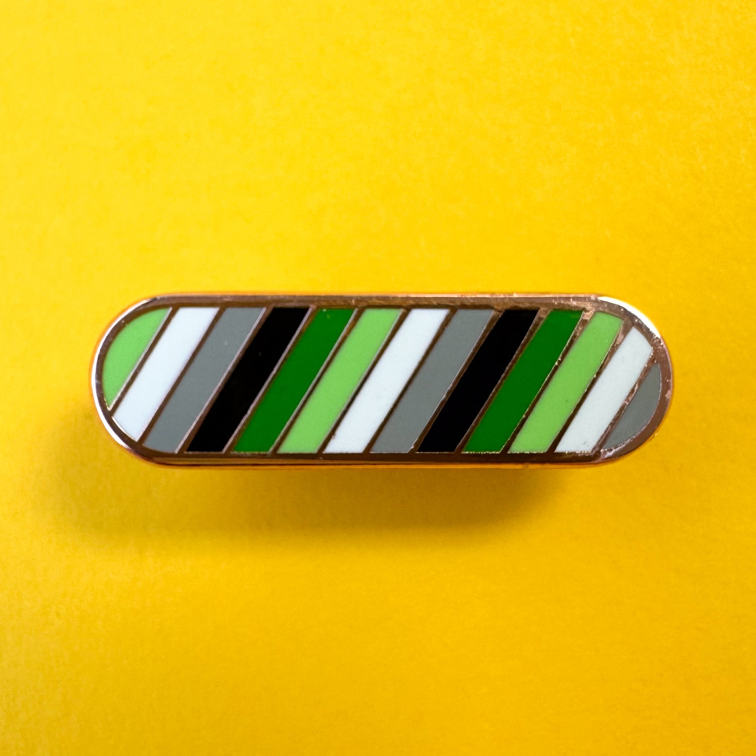 A bandaid shaped pin with diagonal bands of color in the colors of the Aromantic Pride Flag, green, light green, white, grey and black. The pin is on a yellow background.