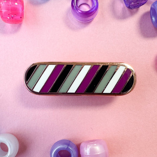 A bandaid shaped pin with diagonal bands of color on it in black, purple, white, and grey representing the asexual or ace flag. The pin is on a light pink background with pony beads around it.