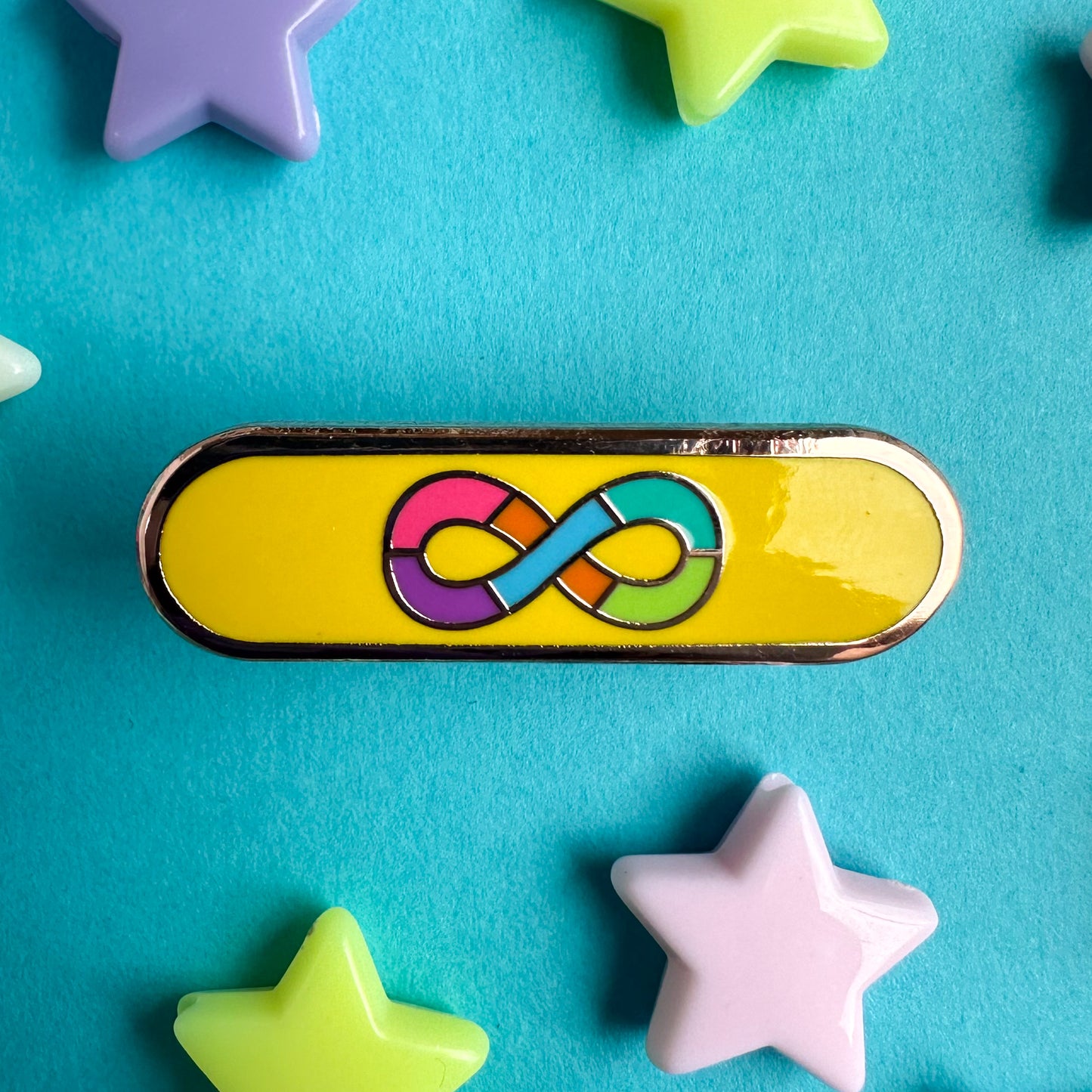 An oval shaped pin with the image of the Autistic Pride Flag on it, a rainbow infinity symbol with a gold background. The pin is on a blue piece of paper with pastel plastic star beads around it. 