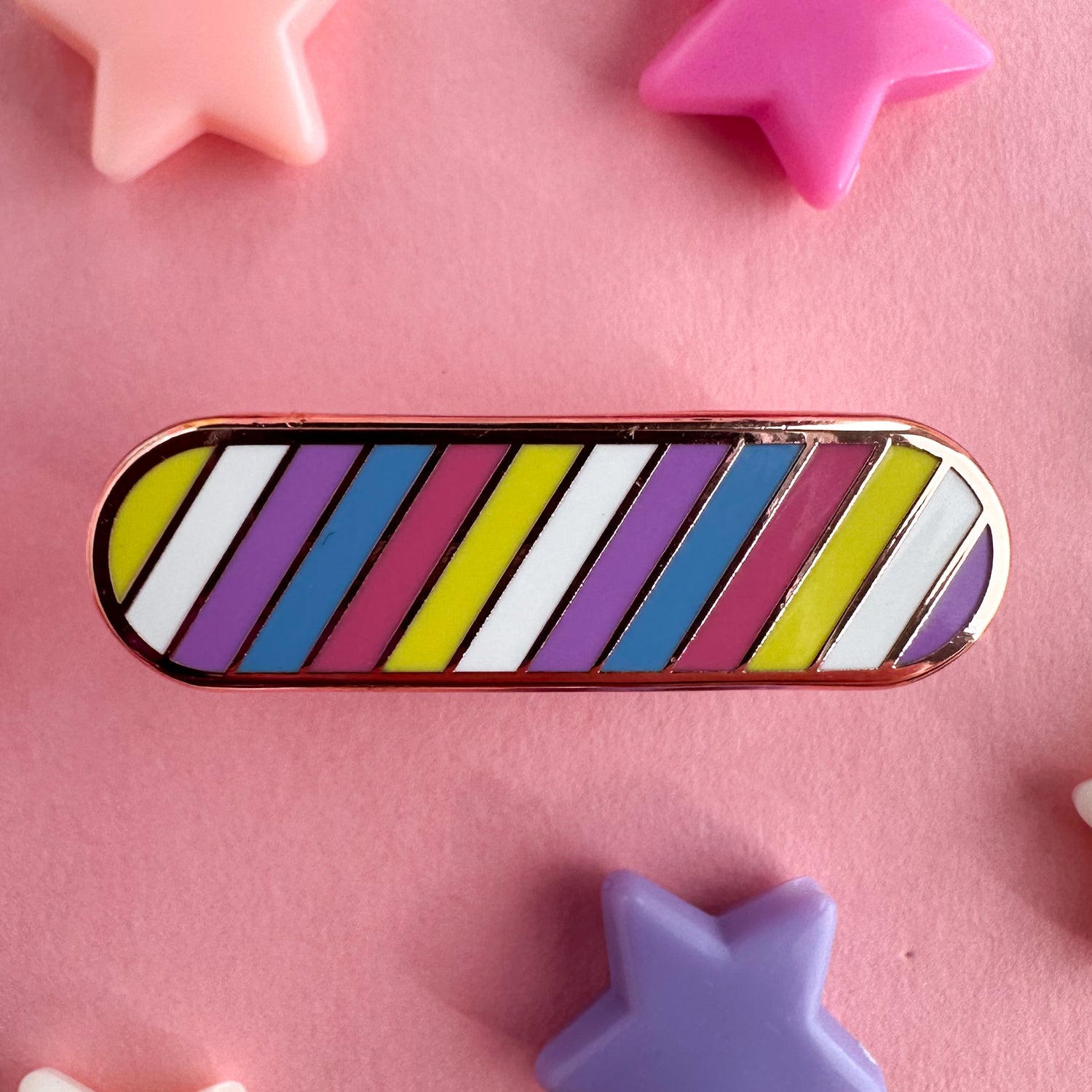 A capsule shaped enamel pin in the colors of the bigender pride flag. The pin is on a pink background with pastel star beads around it. 