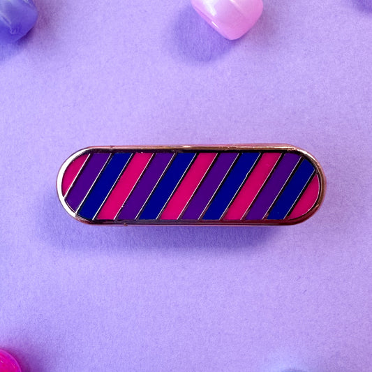 A bandaid shaped enamel pin with diagonal stripes in pink, purple, and blue which are the colors of the Bi Pride Flag. The pin is on a lavender background with pony beads around it.