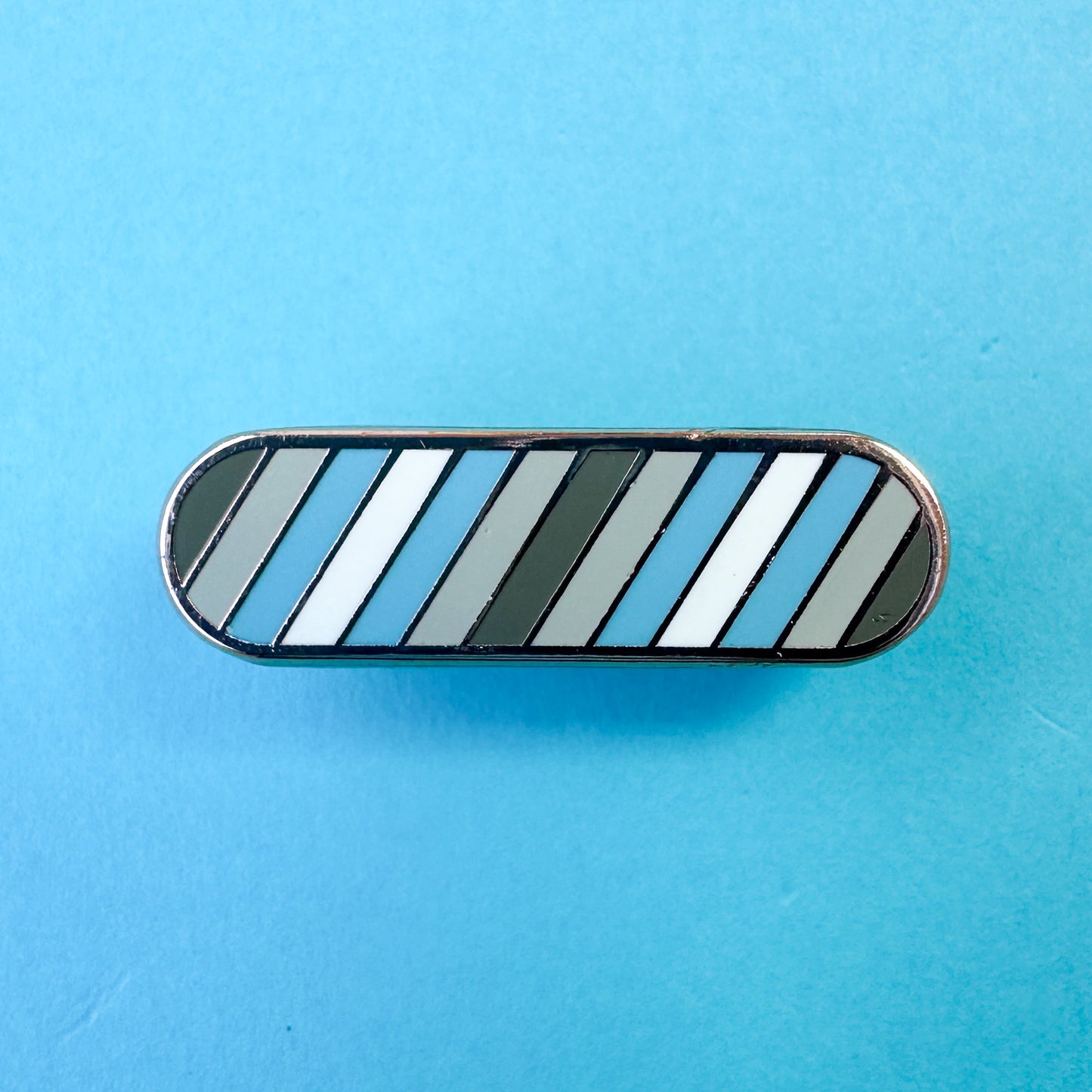 A bandaid shaped pin with diagonal stripes in grey, light grey, light blue, and white, which are the colors of the Demiboy Pride flag. The pin is on a blue background