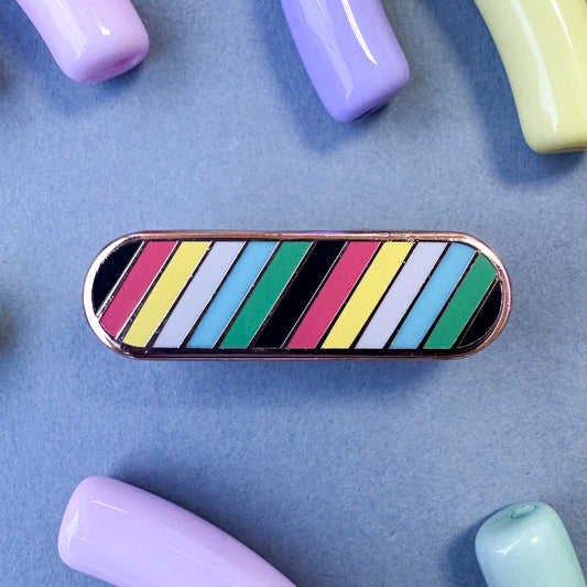 A long oval shaped pin with diagonal stripes of the colors of the Disability Pride flag. The pin is on a grey paper with pastel beads around it.