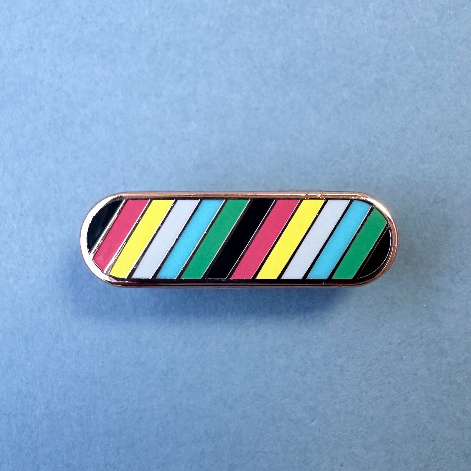 A long oval shaped pin with diagonal stripes of the colors of the Disability Pride flag. The pin is on a grey background.