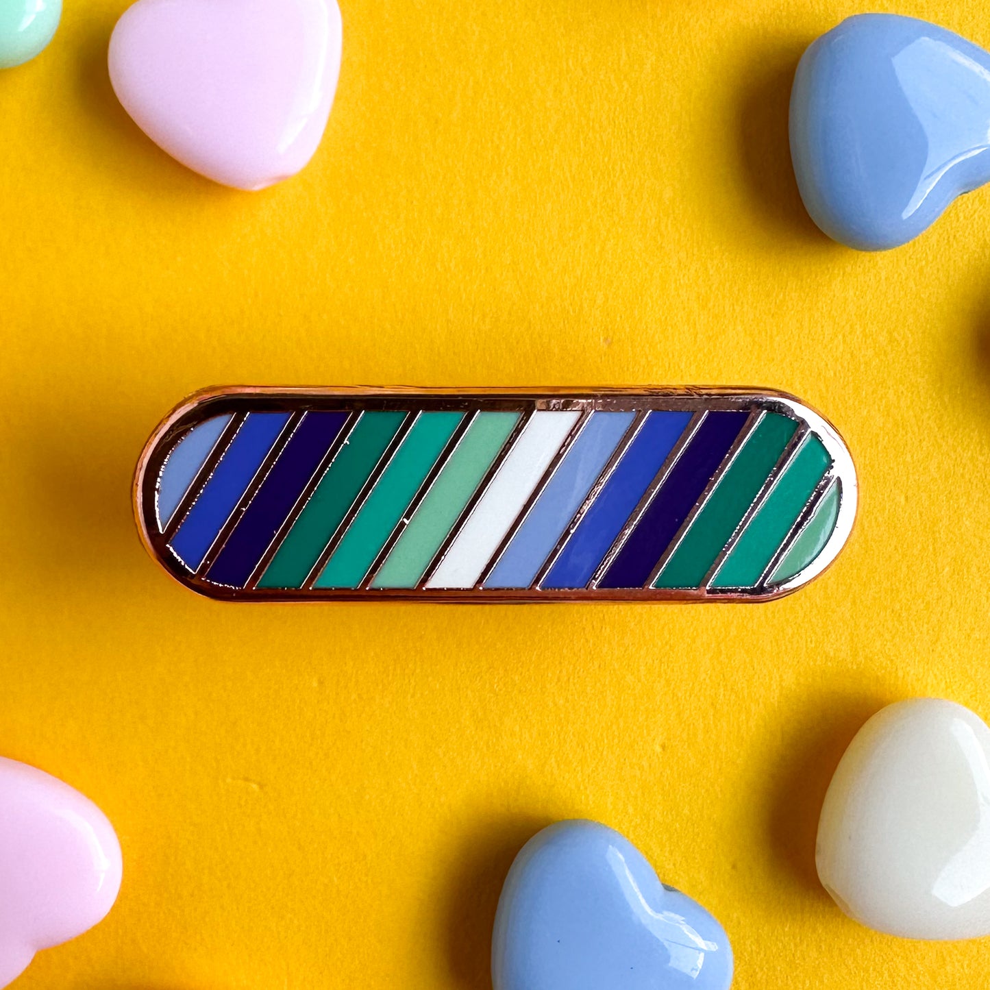 A capsule shaped pin with diagonal stripes in the colors of the gay men flag. The pin is on a yellow background with heart beads around it. 