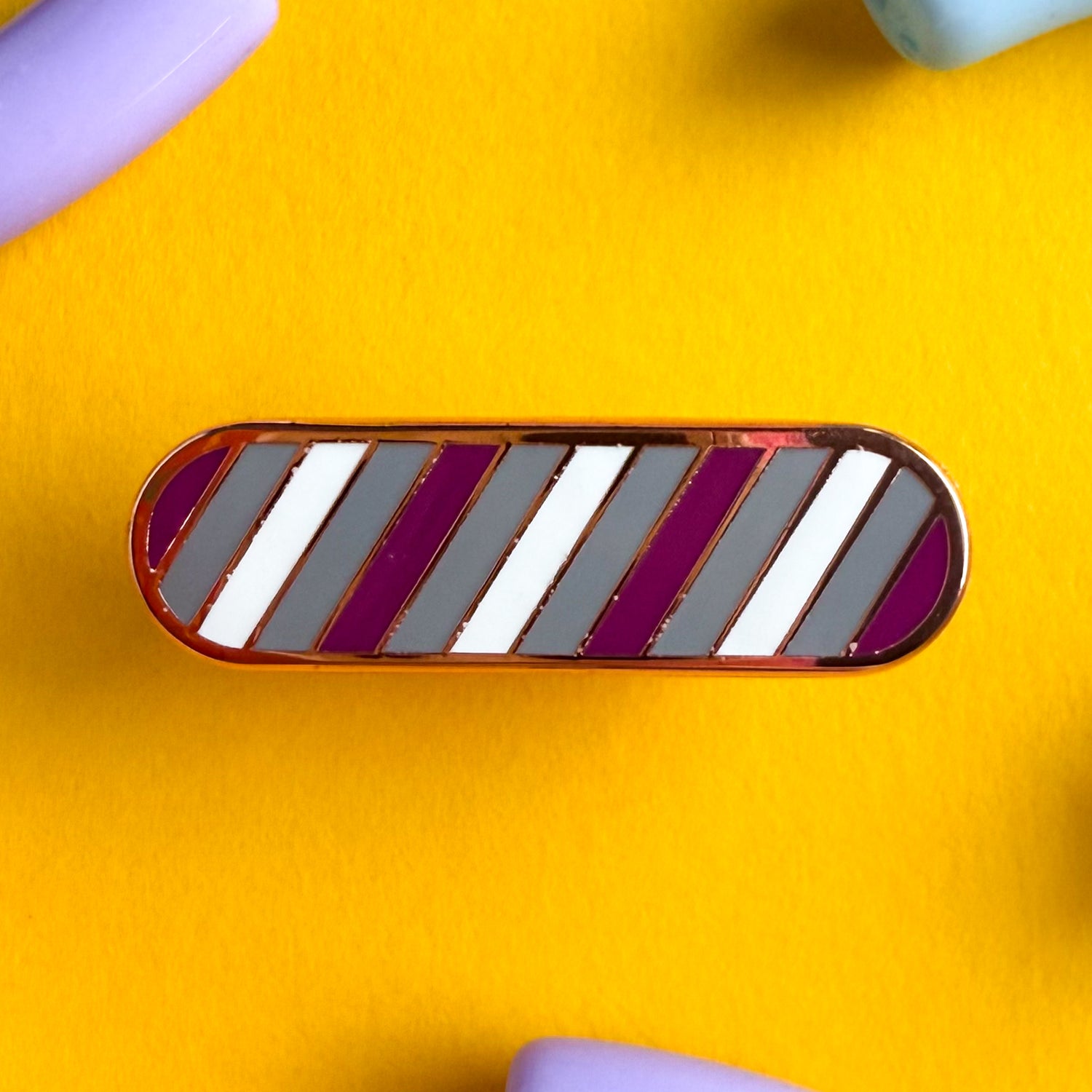 A bandaid shaped pin with diagonal bands of color on it in gray, purple, and white representing the gray asexual or gray ace flag. The pin is on a yellow background.