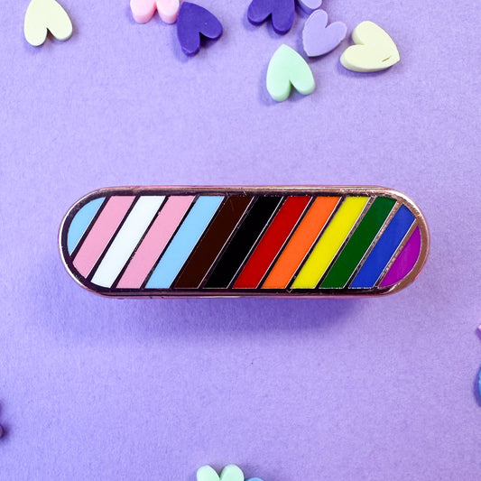 An oval shaped pin with diagonal stripes in the colors of the progress pride flag. The pin is on a lavender background with pastel heart confetti around. 