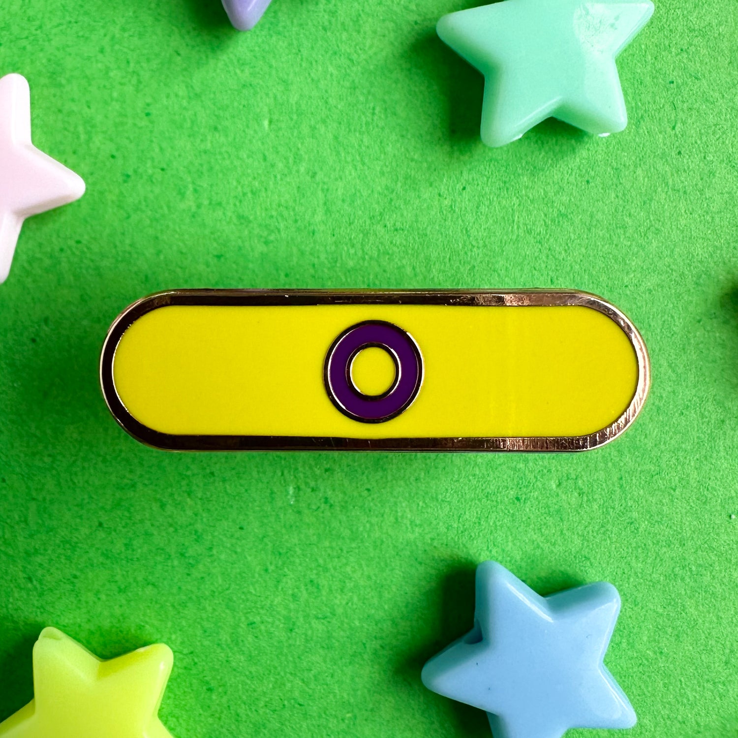 A capsule shaped pin that has a purple circle on a yellow background to represent the Intersex Pride Flag. The pin is on a green paper background with pastel star beads around it. 