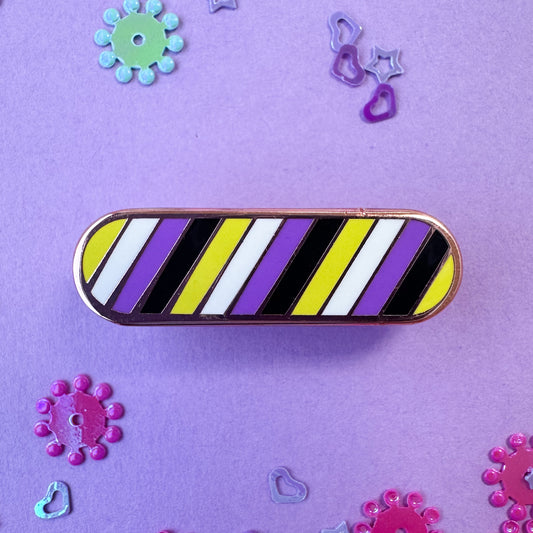 An oval shaped pin with stripes of the colors of the Nonbinary flag. The pin is on a purple background with pink and blue confetti around it.