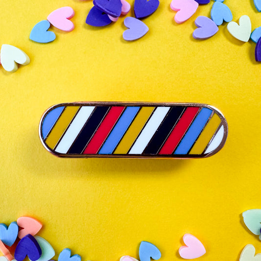 A capsule pin with diagonal stripes in the color of the Poly pride flag. The pin is on a yellow background with heart confetti around it. 