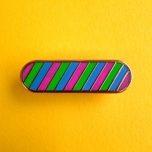 A capsule shaped enamel pin in the colors of the Polysexual pride flag which are pink, green, and blue. The pin is on a yellow paper background.
