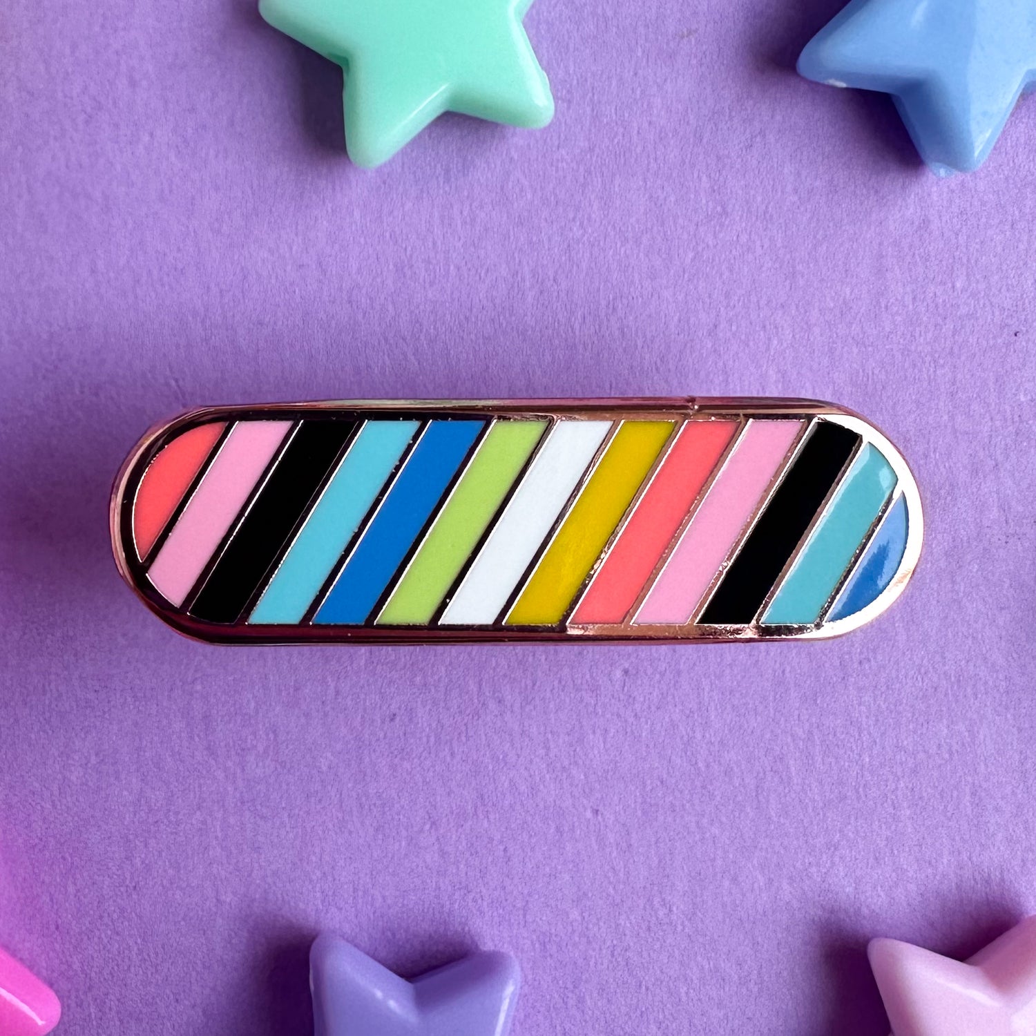 An enamel pin shaped like a bandaid in the colors of the Queer Pride Flag with diagonal stripes. The pin is on a purple background with plastic star beads around it. 