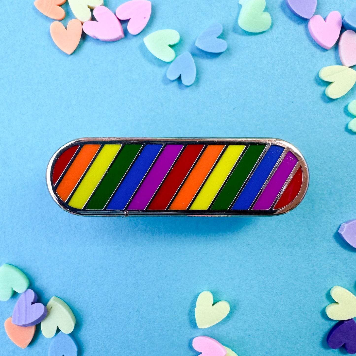 A capsule shaped enamel pin with rainbow stripes in diagonal lines. The pin is on a blue background with confetti hearts around. 