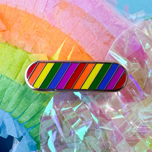 An oval pin with diagonal stripes in the colors of the rainbow. The pin is on top of a pastel rainbow piñata. 