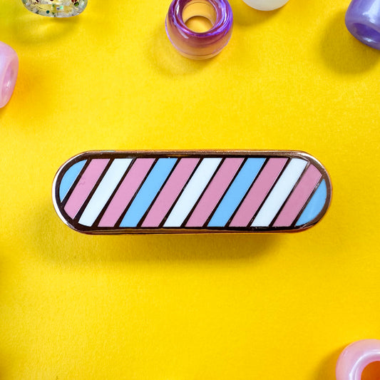 An oval shaped pin in the colors of the Trans pride flag light blue, light pink, and white. The pin is on a yellow background with pony beads around it.