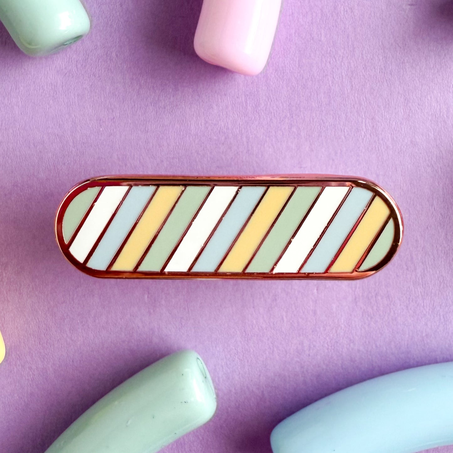 A bandaid shaped pin with diagonal bands of pastel color on it in blue, yellow, green, and white representing the unlabeled flag. The pin is on a purple background.