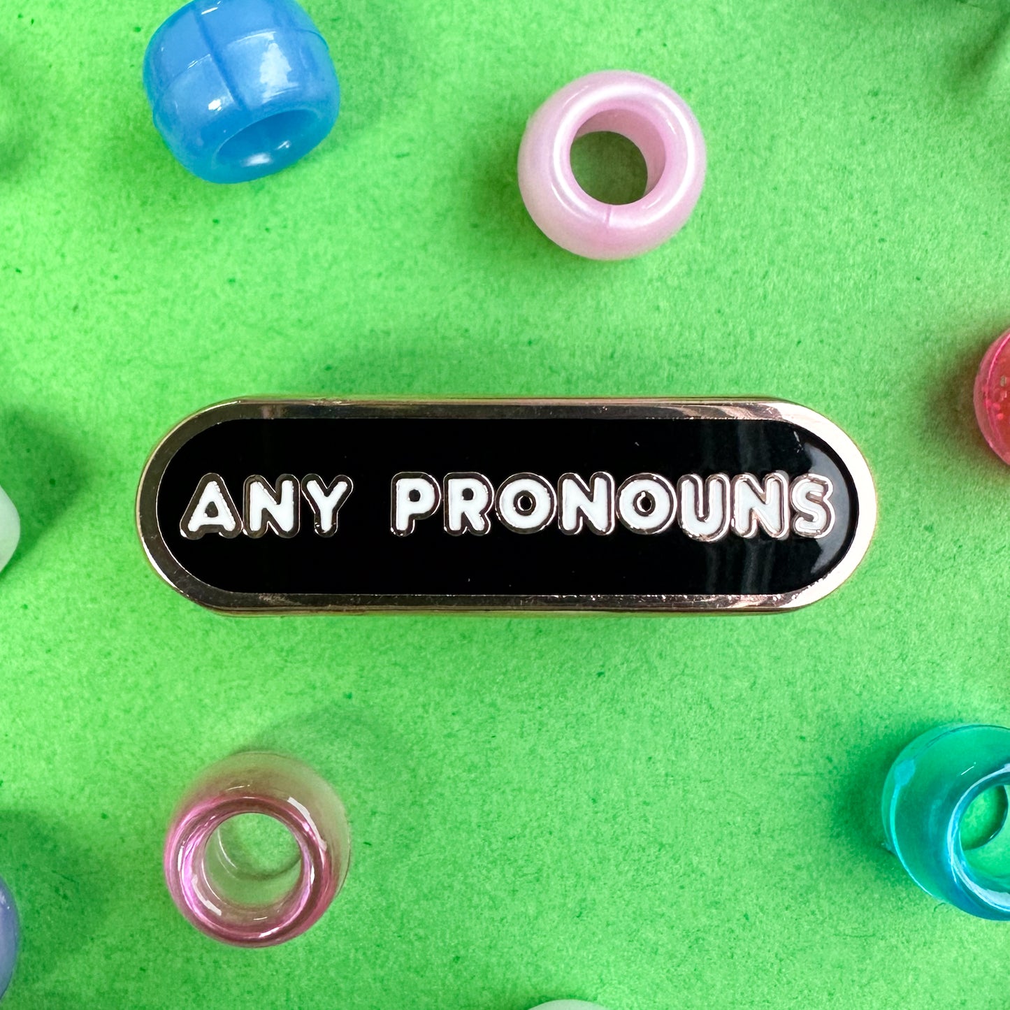 A bandaid shaped pins with white letters that read "any pronouns" on a black background. The pin is on a green background with pony beads around it. 