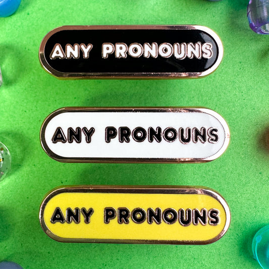 Bandaid shaped pins with the words "any pronouns" on them, the top pin is black, the middle is white and the last one is yellow the pins are on a green background with beads around it.