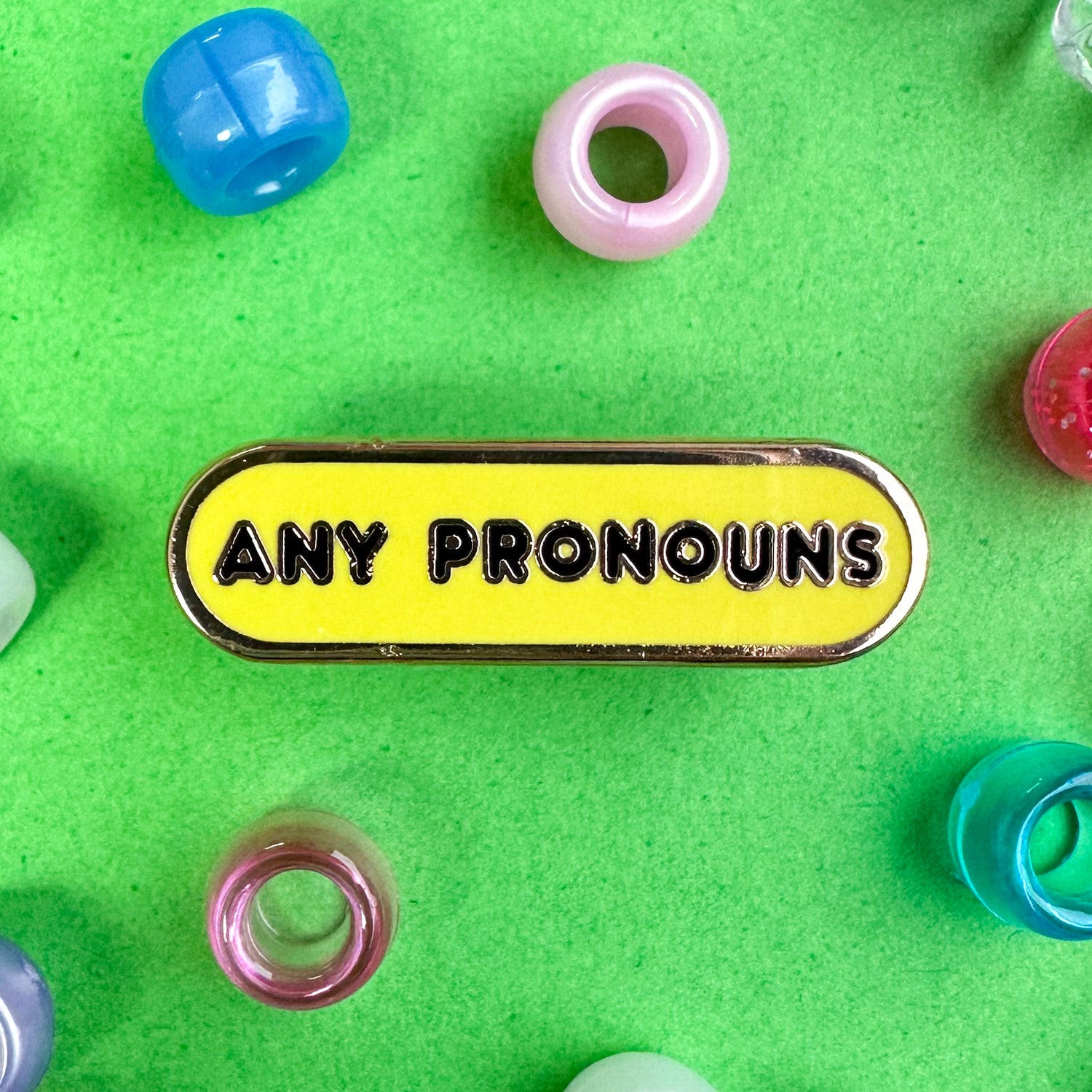 A bandaid shaped enamel pin with black letters that read "Any pronouns" with a yellow background. The pin is on a green background with pony beads strewn around it. 