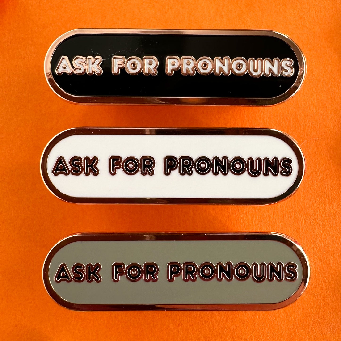 Three enamel pins that all read "Ask For Pronouns" in bubble letters on a capsule shaped background. The top pin has a black background, the middle white, the bottom grey. The pins are on an orange background.