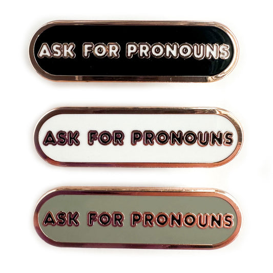 Three enamel pins that all read "Ask For Pronouns" in bubble letters on a capsule shaped background. The top pin has a black background, the middle white, the bottom grey.