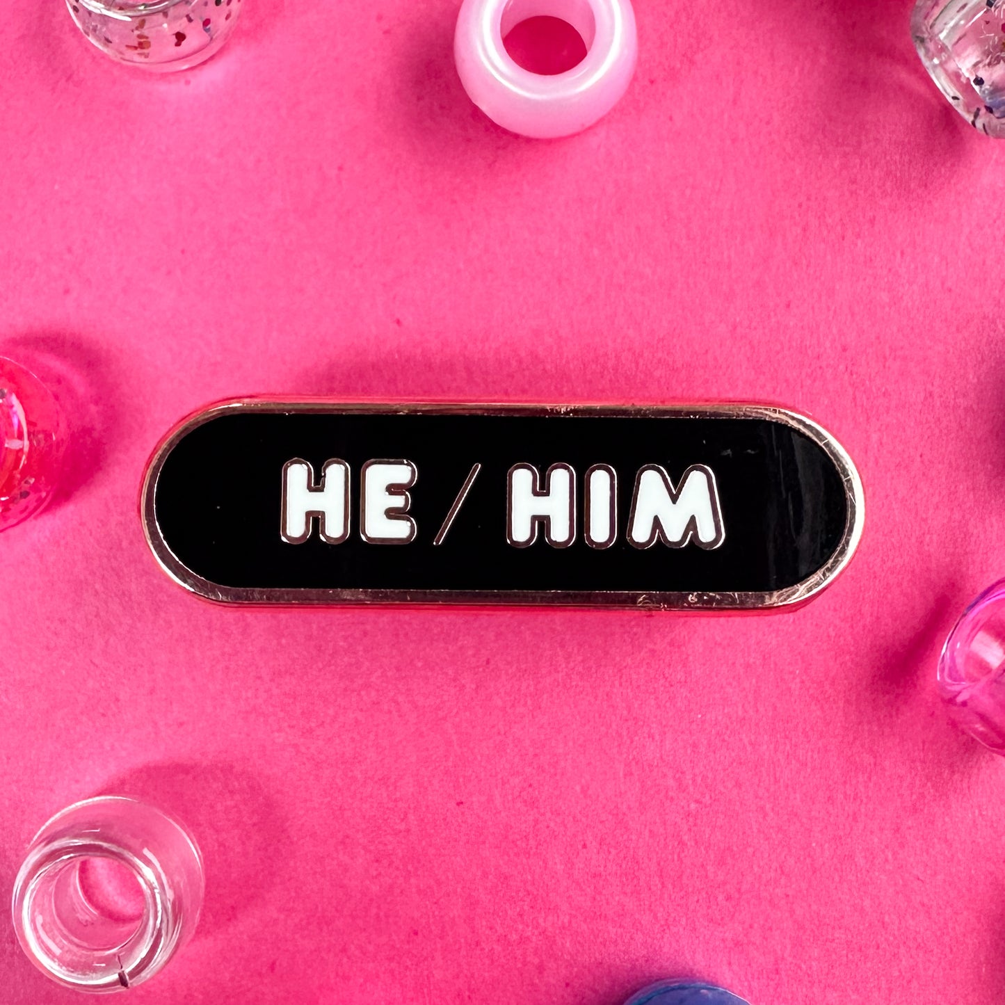 A black capsule pin with white bubble letters that read "He/Him". The pin is on a hot pink background with pony beads around it. 