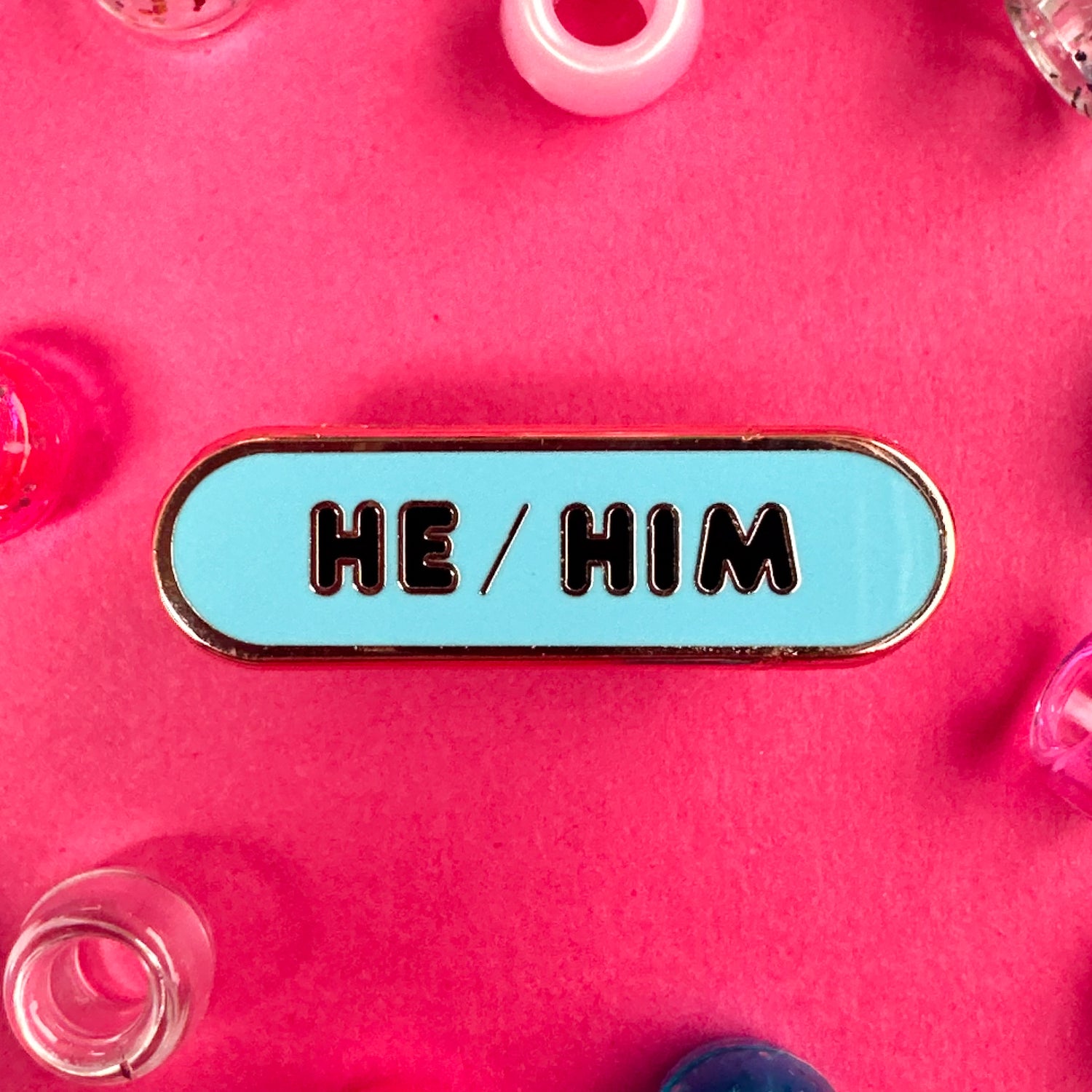 A blue oval shaped pin with the words "He/Him" in rounded letters. The pin is on a hot background with pony beads around it. 