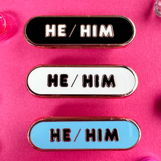Three pronoun pins that read "He/Him" they are oval shaped pins. The pins are on a hot pink background with beads around it. 
