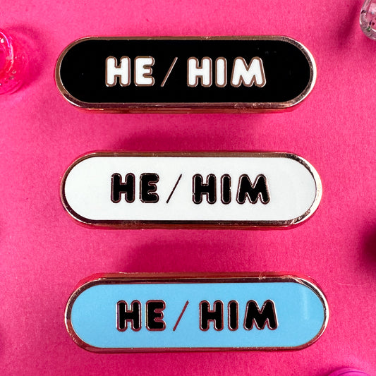 Three pronoun pins that read "He/Him" they are oval shaped pins. The pins are on a hot pink background with beads around it.
