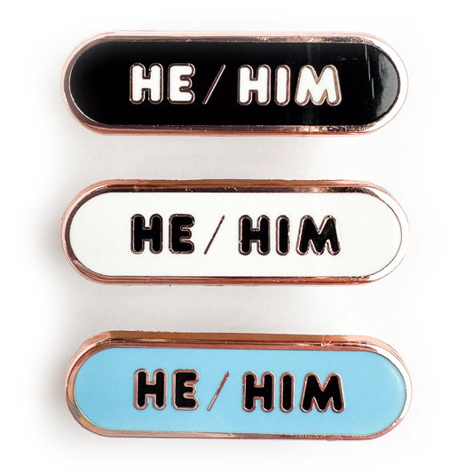 Three capsule shaped pins that all have the words "He/Him" in bubble letters. One of the pins is black, one is white and one is light blue. 