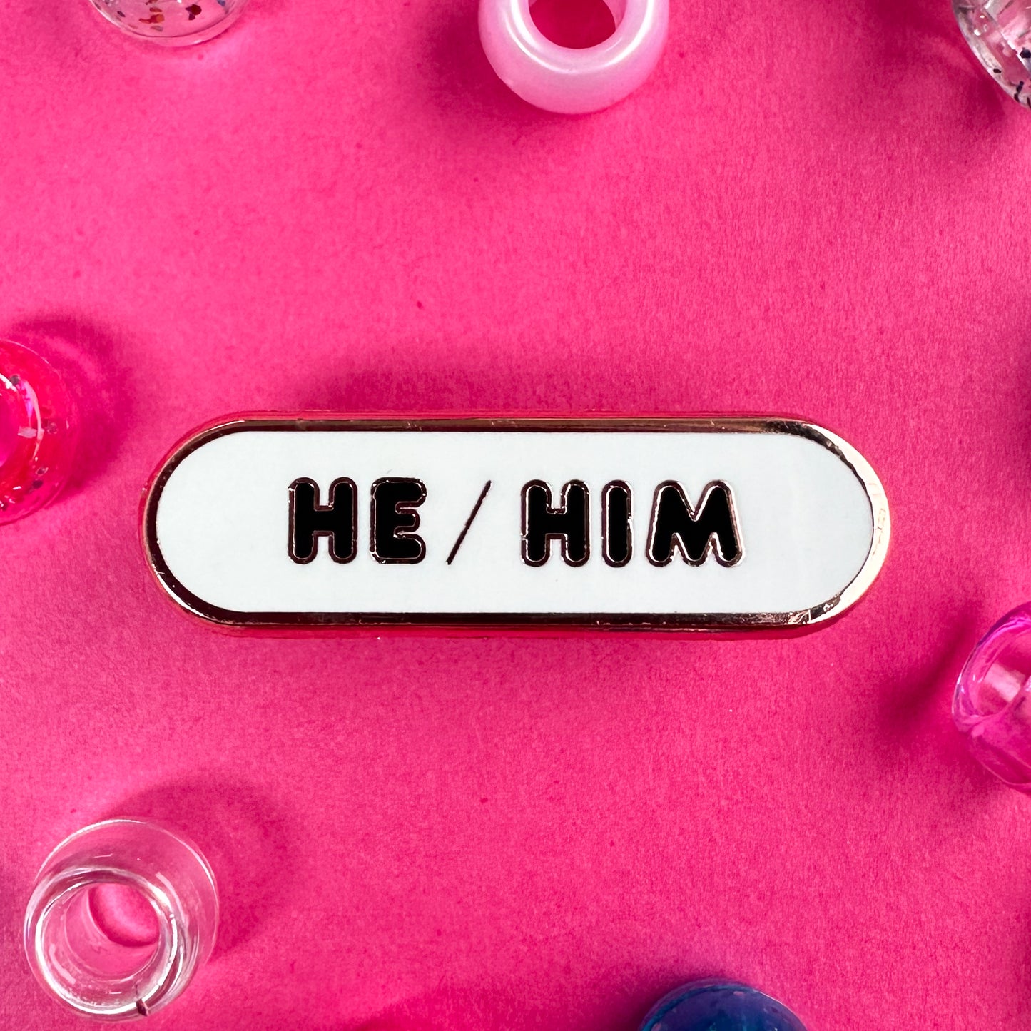 A white capsule pin with bubble letters that read "He/Him". The pin is on a hot pink background with pony beads around it. 