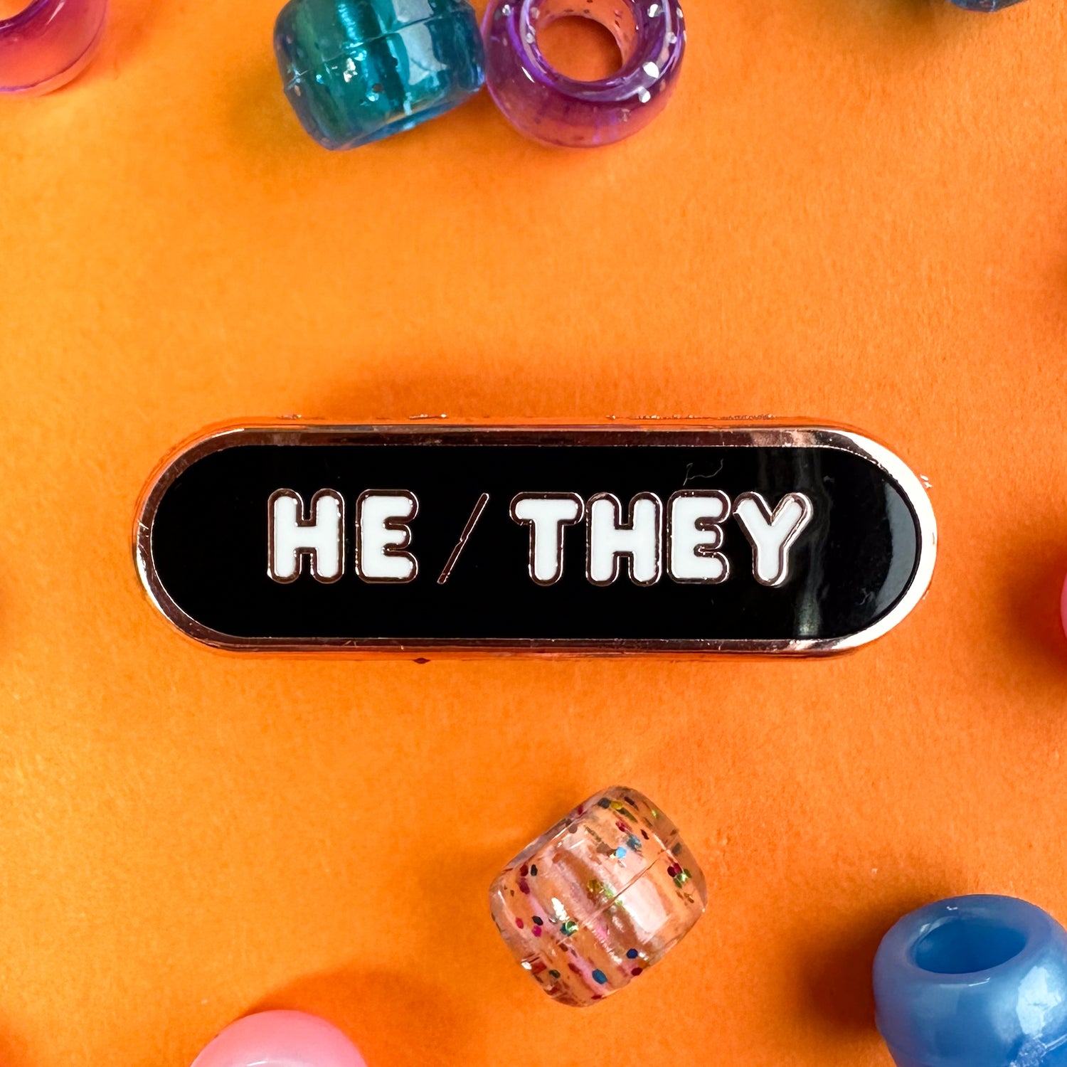 A black capsule shaped pin with white bubble letters that spell "He/They" to share your pronouns. The pin is on an orange background with pony beads around.