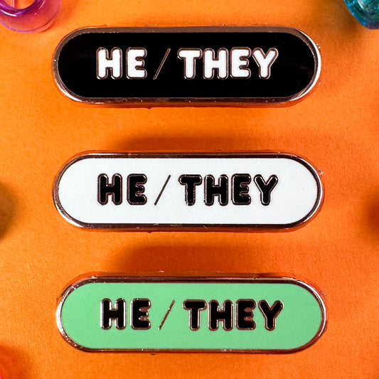 Three different oval shaped pins that read "He/They" one is black, one is white, and one is mint green. The pins are on an orange background with pony beads.