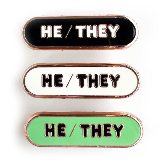 Three capsule shaped pins that all have words that read "He/They" one of the pins is black, one is white, and one is mint green.