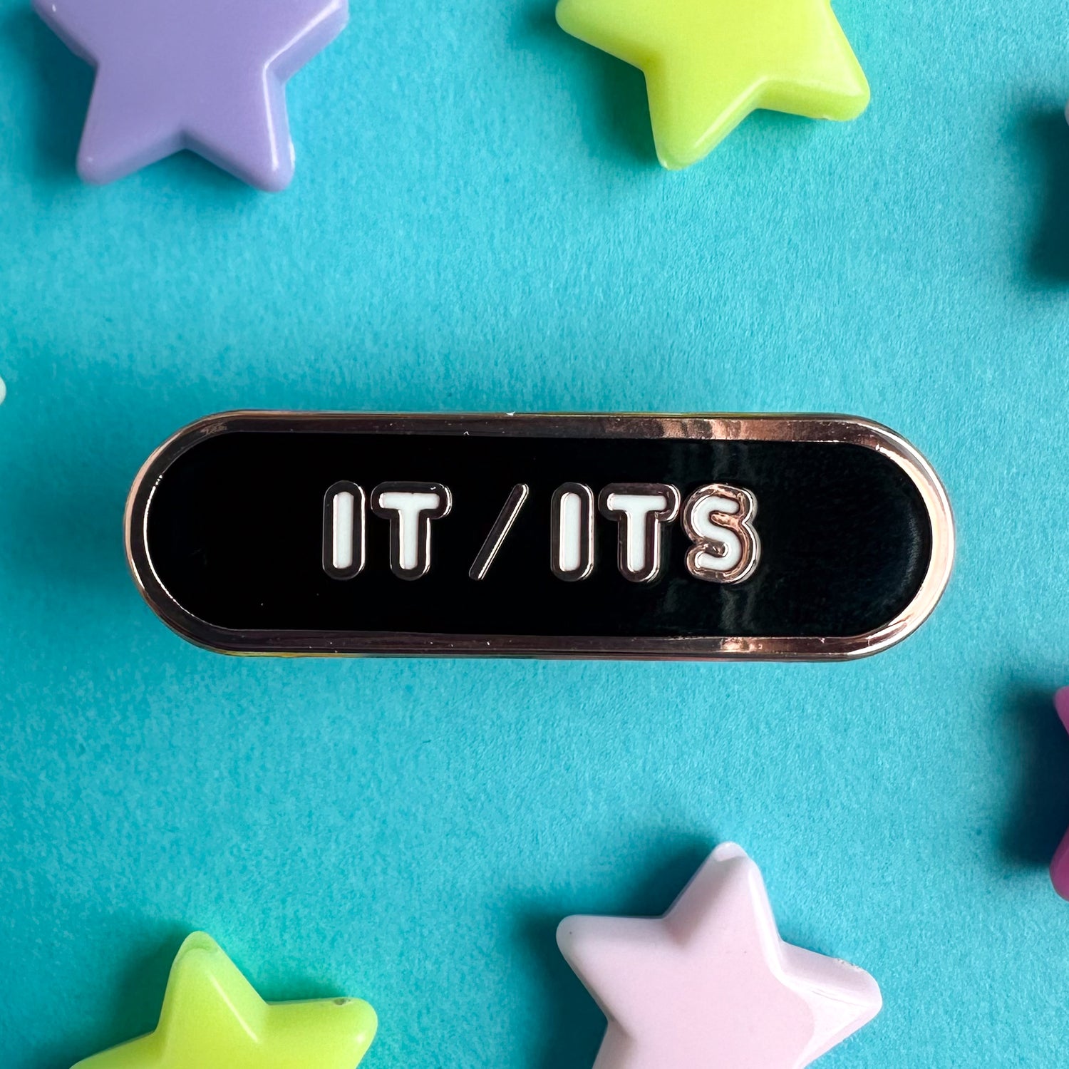 A black capsule shaped enamel pin with white bubble letters that read "It/Its". The pin is sitting on a blue piece of paper with plastic star beads around it. 