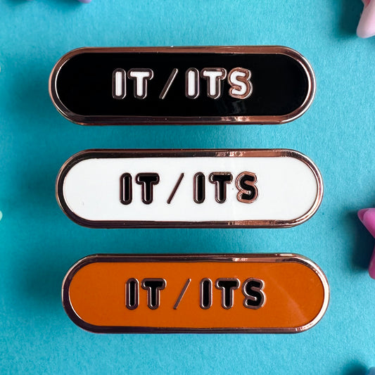 Three enamel pins that all read "It/Its" the top pin is black, the middle is white, and the bottom is orange. All of the pins are capsule shaped. The pins are on a blue background.