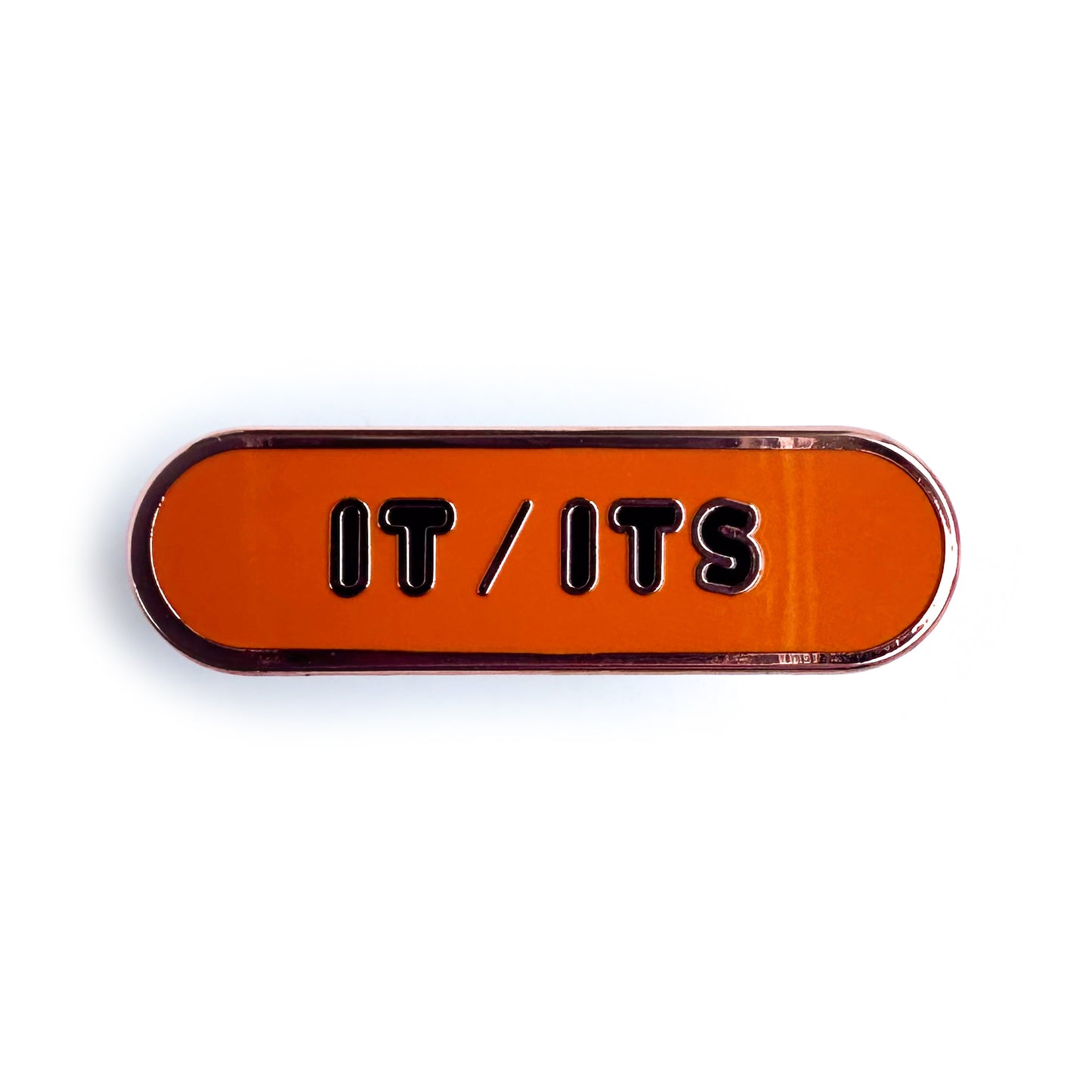 An orange capsule shaped enamel pin with the words "It/Its" on it in black bubble letters.