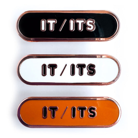 Three enamel pins that all read "It/Its" the top pin is black, the middle is white, and the bottom is orange. All of the pins are capsule shaped. 