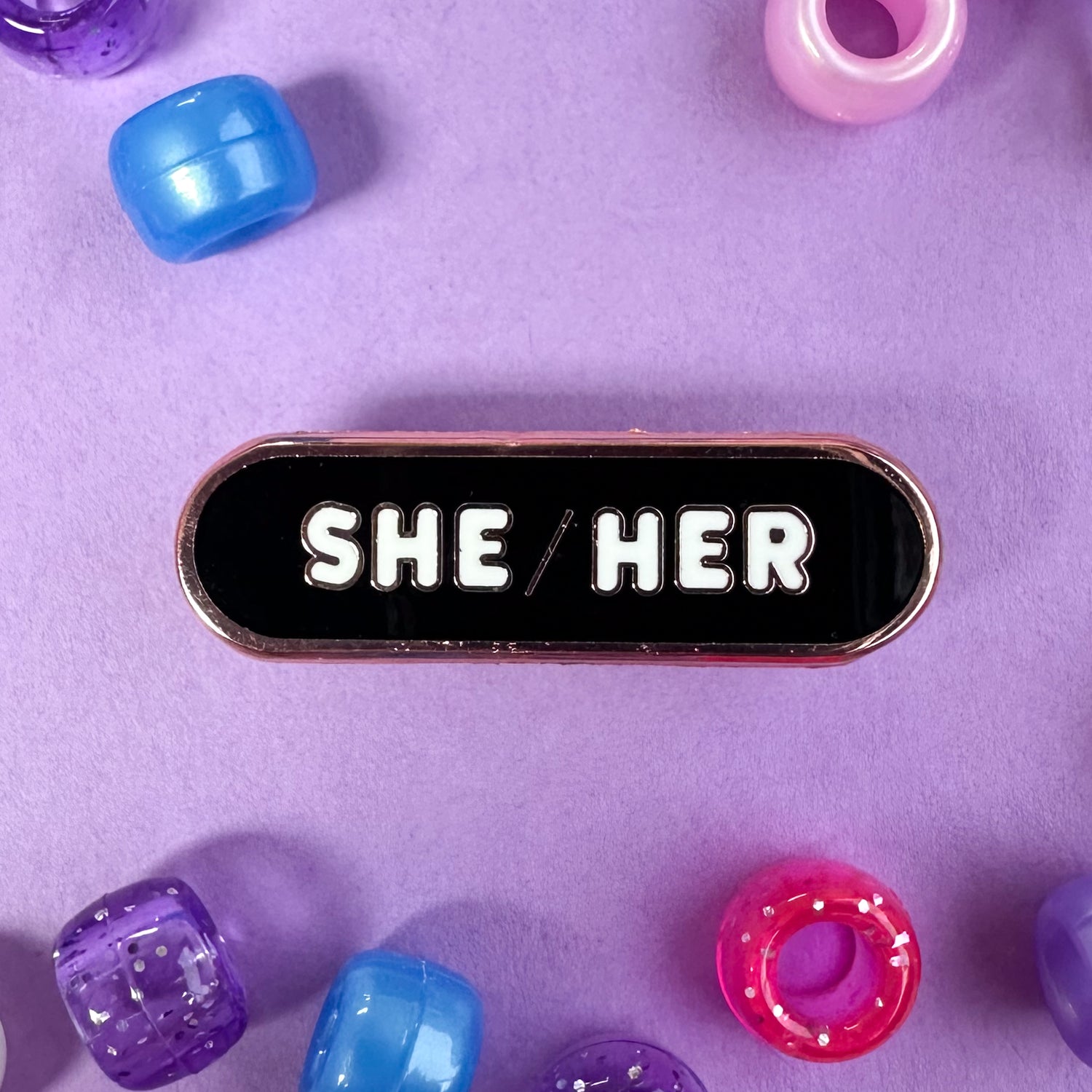 A capsule shaped pin with the pronouns "She/Her" on it. The pin is on a lavender background with blue, purple, and pink pony beads scattered around it. 