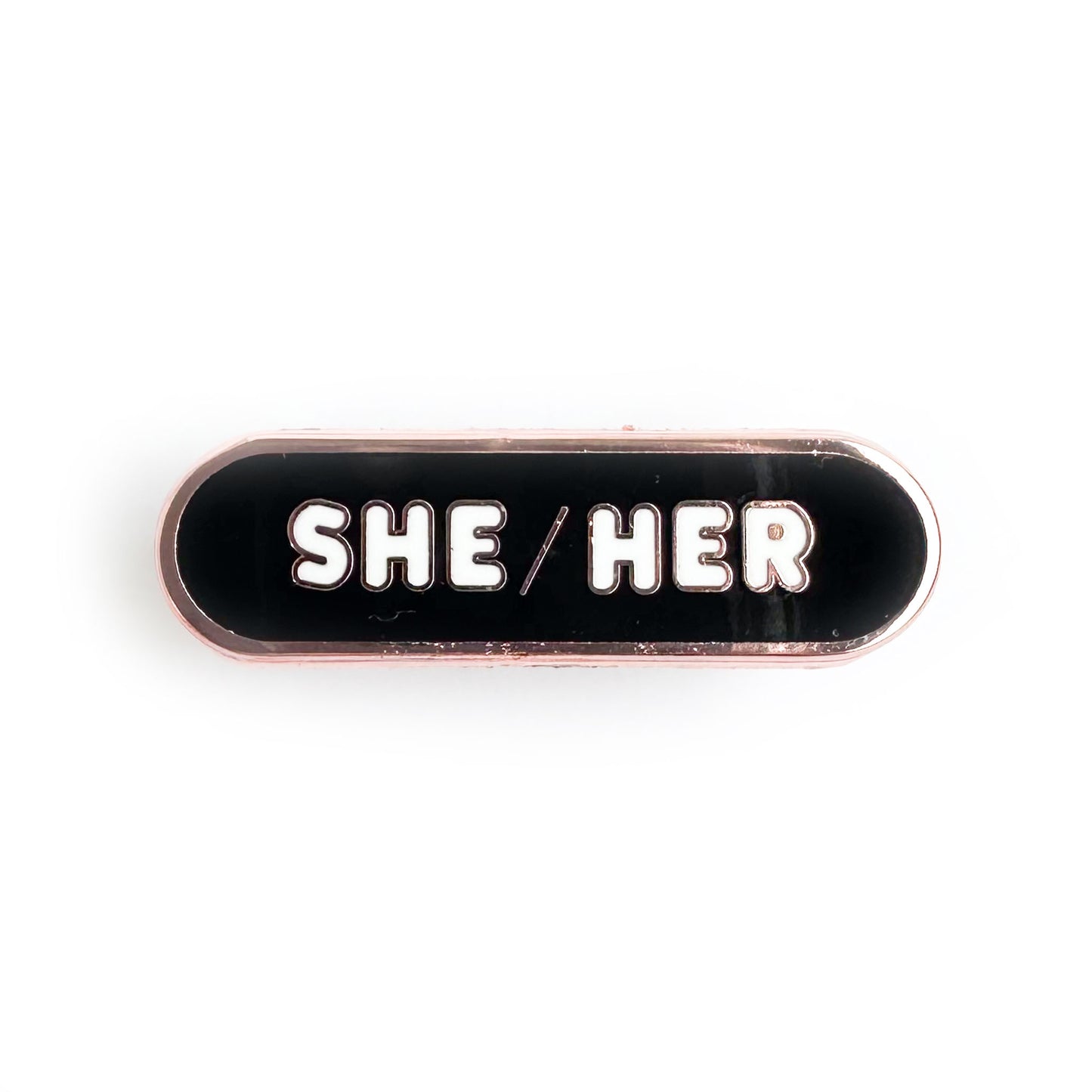 A capsule shaped pin with a black background and white letters that spell the pronouns "She/Her" on it. 