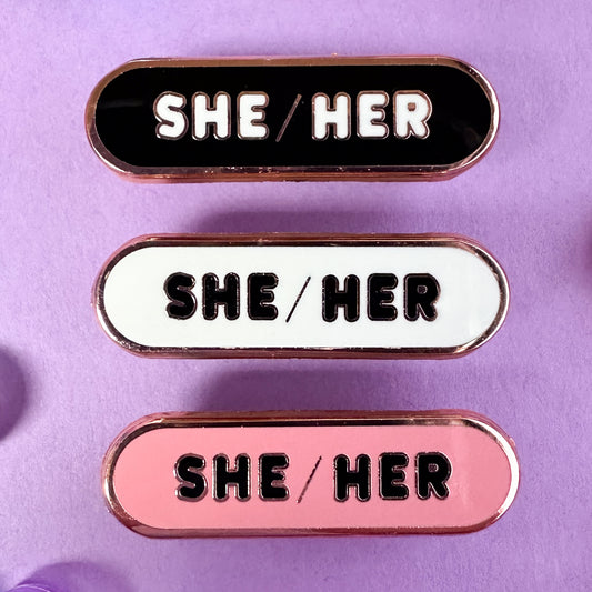 Three oval shaped pins with the words "She/Her" on them, one pin is black, one is white, and one is pink. The pins are on a lavender background. 