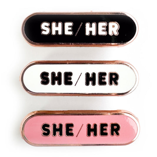 Three capsule shaped pins that read "She/Her' one is black, one is white, and one is pink.