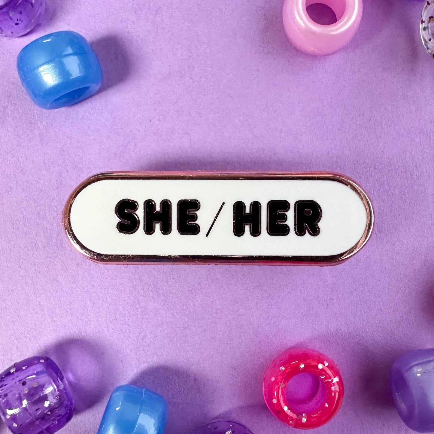 A capsule shaped pin with the pronouns "She/Her" on it. The pin is on a lavender background with pink, purple, and blue pony beads scattered around. 