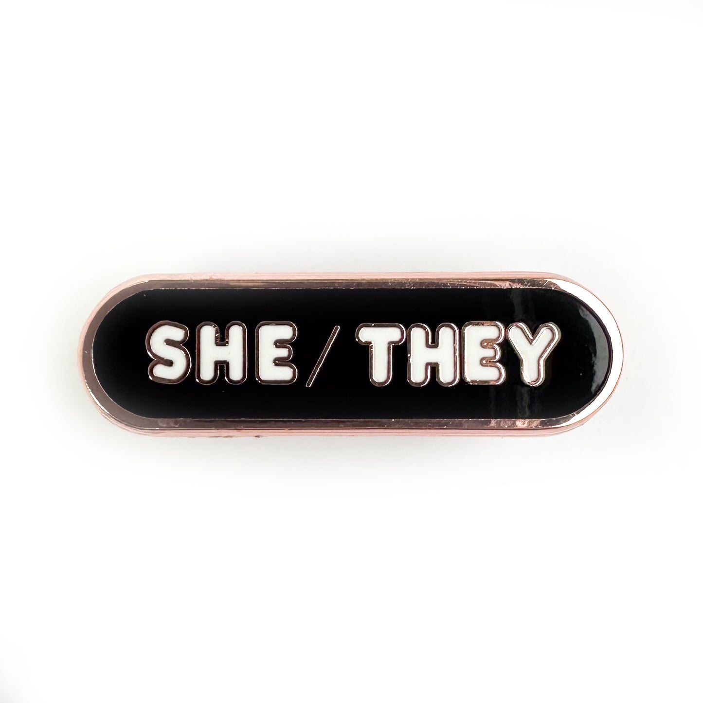 A pin shaped like a bandaid with white letters that read "She/They" on a black background. 