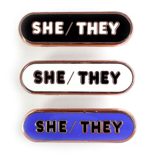 Three capsule shaped pins that read "She/They" one is in black, one is in white, and one is in indigo.
